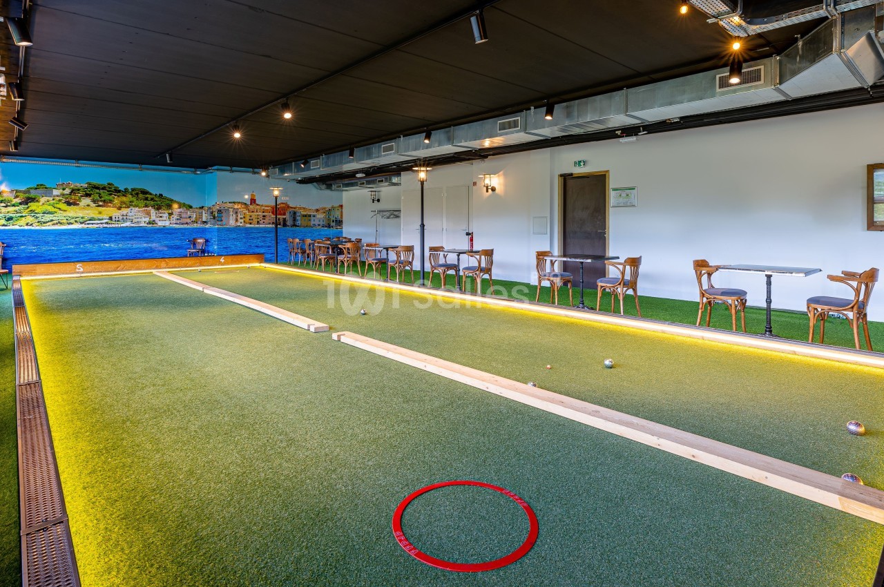 Terrain de pétanque intérieur avec éclairage chaleureux, tables et chaises en arrière-plan, décor mural méditerranéen.