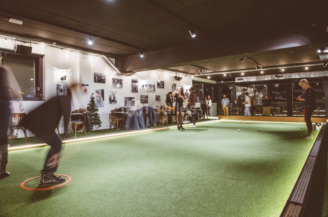 Personnes jouant à la pétanque en intérieur sur un terrain éclairé, avec des photos accrochées aux murs.