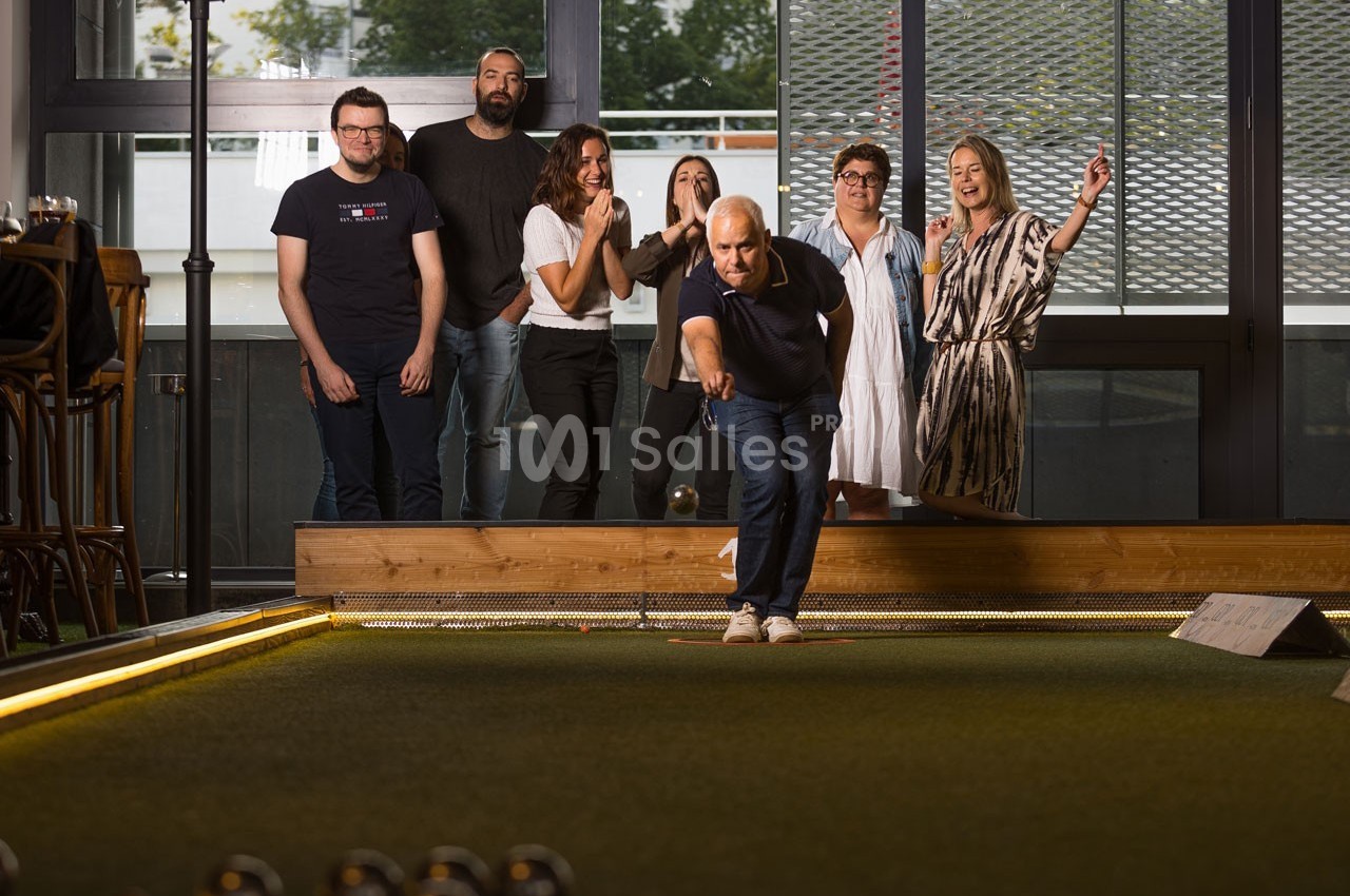 Un groupe de personnes regarde un homme jouer à la pétanque dans un espace intérieur lumineux.