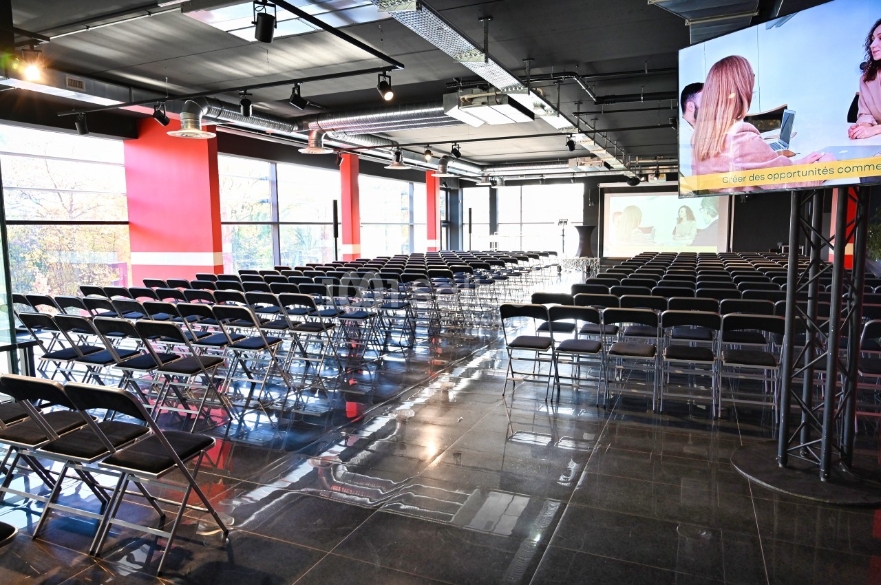 Salle de conférence moderne avec des rangées de chaises vides, écran de projection et baies vitrées.