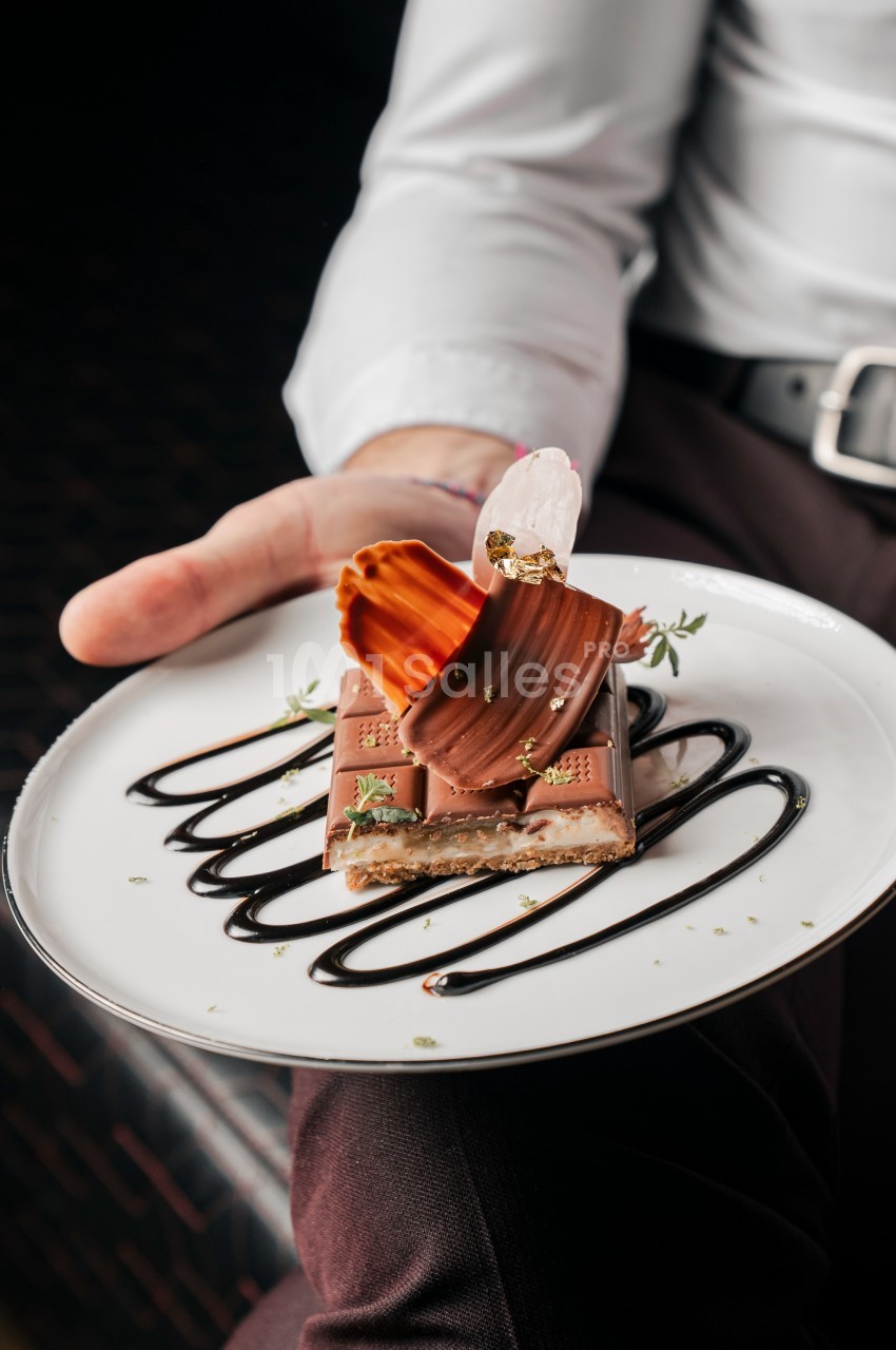 Assiette de dessert avec chocolat, décorations en caramel et sauce au chocolat, tenue par une personne en chemise blanche.