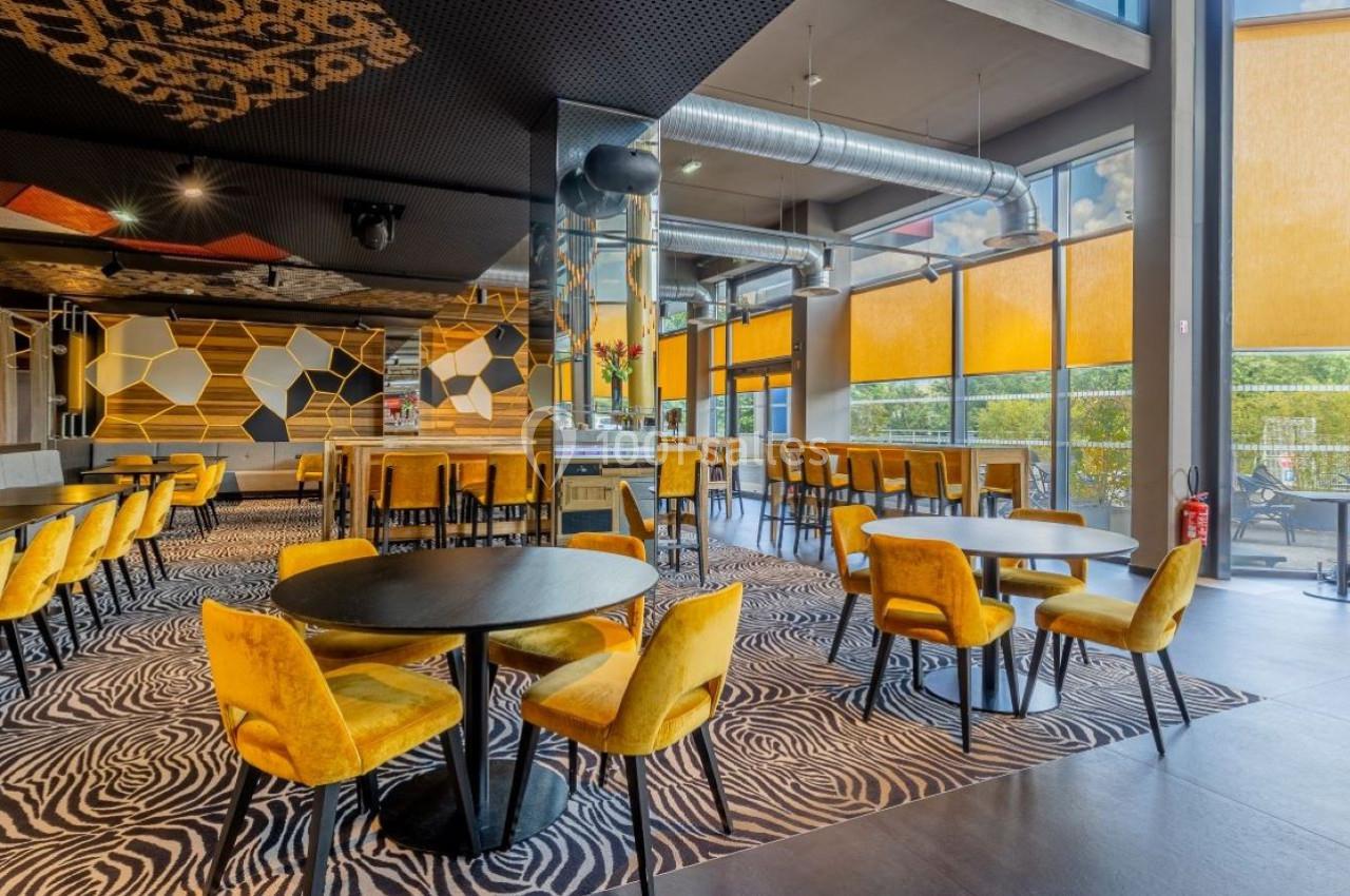 Salle de restaurant moderne avec tables noires, chaises jaunes, tapis à motifs et grandes baies vitrées avec stores.
