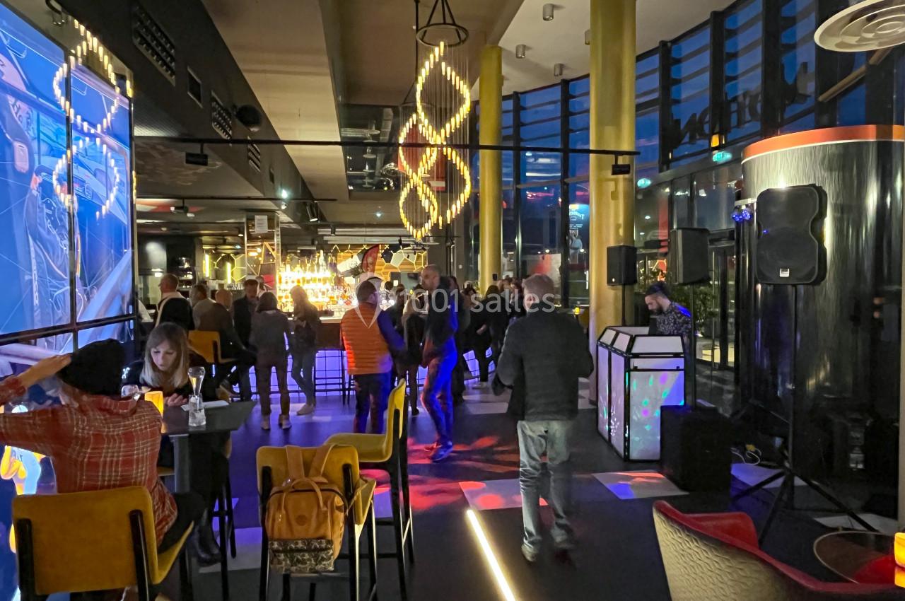 Personnes discutant et dansant dans un bar moderne avec éclairage coloré et grandes baies vitrées en soirée.