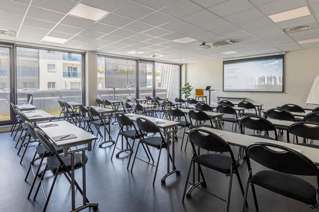 Salle de formation lumineuse avec des rangées de tables et chaises, écran de projection et grandes fenêtres.