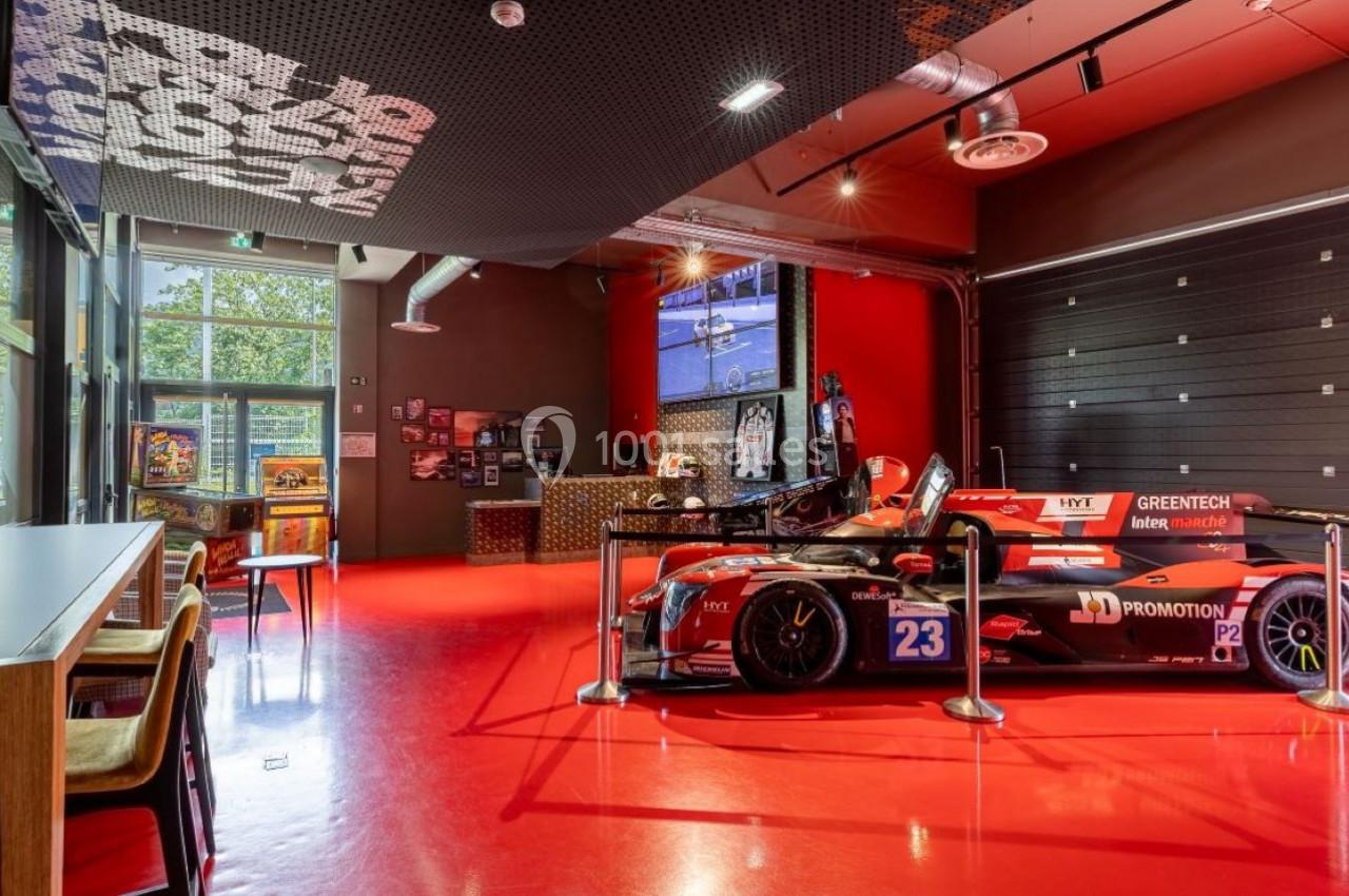 Voiture de course rouge exposée dans une salle moderne avec sol rouge, mobilier en bois et baies vitrées.