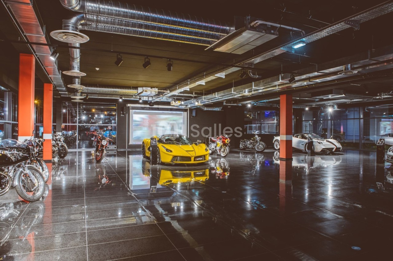 Salle d'exposition moderne avec voitures de sport, motos et éclairage industriel sur sol brillant.