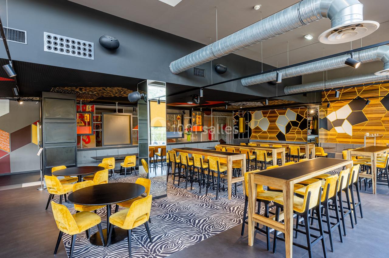 Salle moderne avec tables hautes et basses, chaises jaunes, décor géométrique coloré et conduits apparents au plafond.