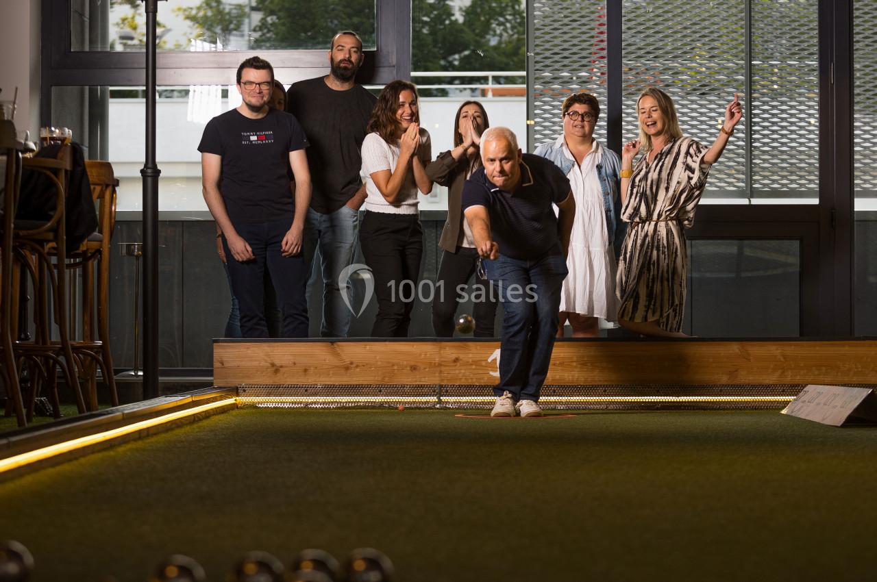 Un groupe de personnes regarde un homme lancer une boule sur un terrain de pétanque en intérieur.