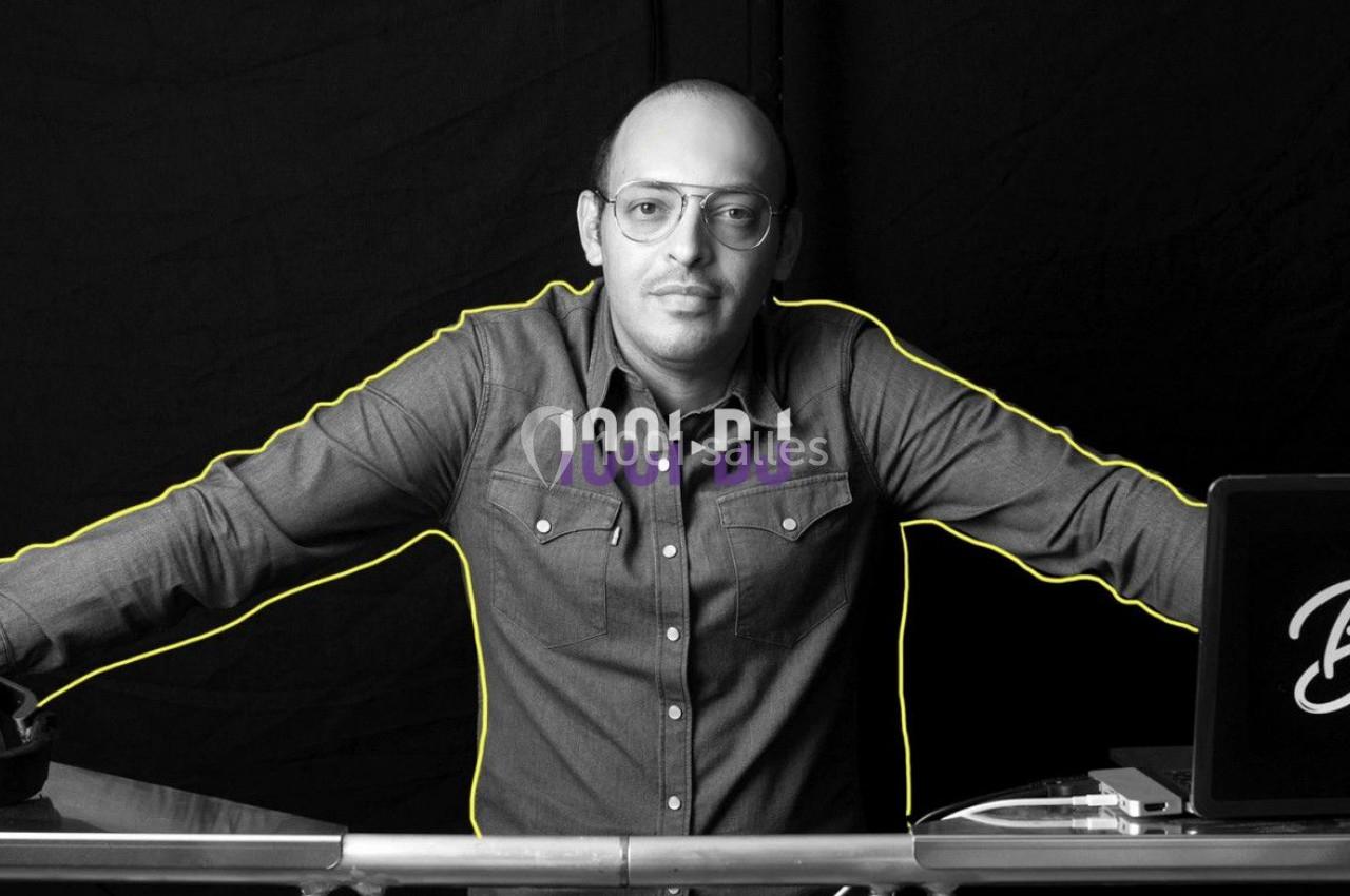 Un DJ debout derrière une table avec un casque audio et un ordinateur portable, sur fond noir.