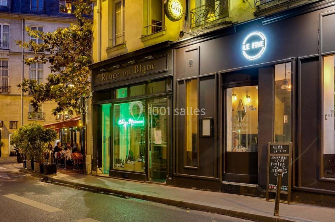 Façade de restaurants et bars éclairés en soirée dans une rue calme avec quelques tables en terrasse.