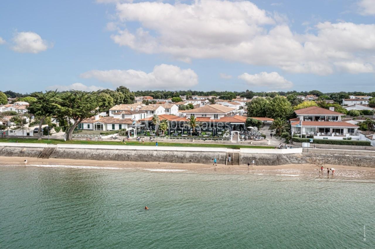 Vue aérienne d'une plage bordée de maisons blanches et végétation, avec des baigneurs au bord de l'eau.