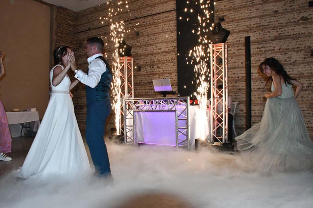Un couple danse au milieu d'une piste entourée de fumée et d'effets pyrotechniques, avec une DJ en arrière-plan.