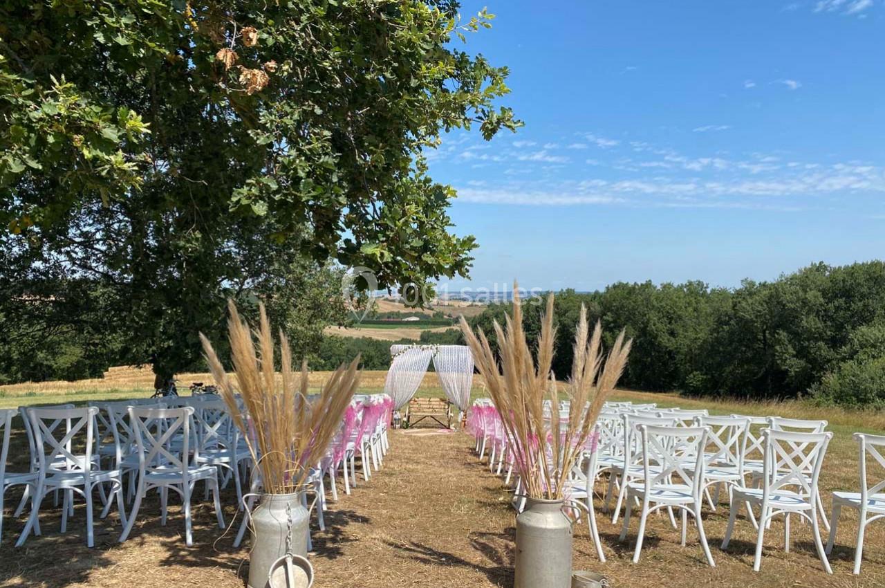 Chaises blanches alignées en extérieur sous un arbre, décorations florales et arche au fond, vue sur la campagne.