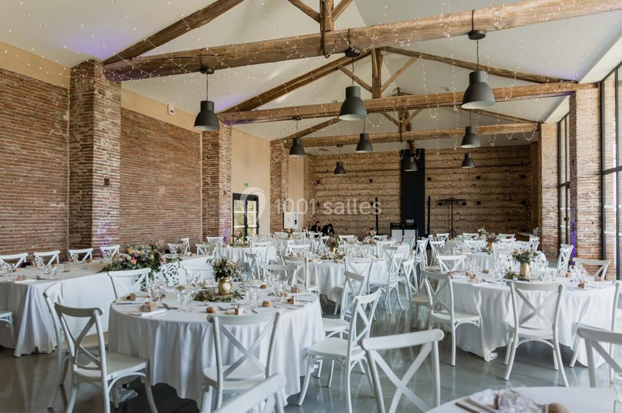 Salle de réception lumineuse avec murs en briques, poutres apparentes et tables rondes décorées pour un événement.
