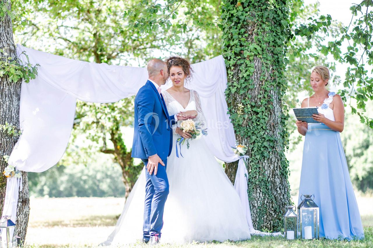 Un couple en tenue de mariage se tient devant un drap blanc suspendu entre des arbres, accompagné d'une femme lisant un…