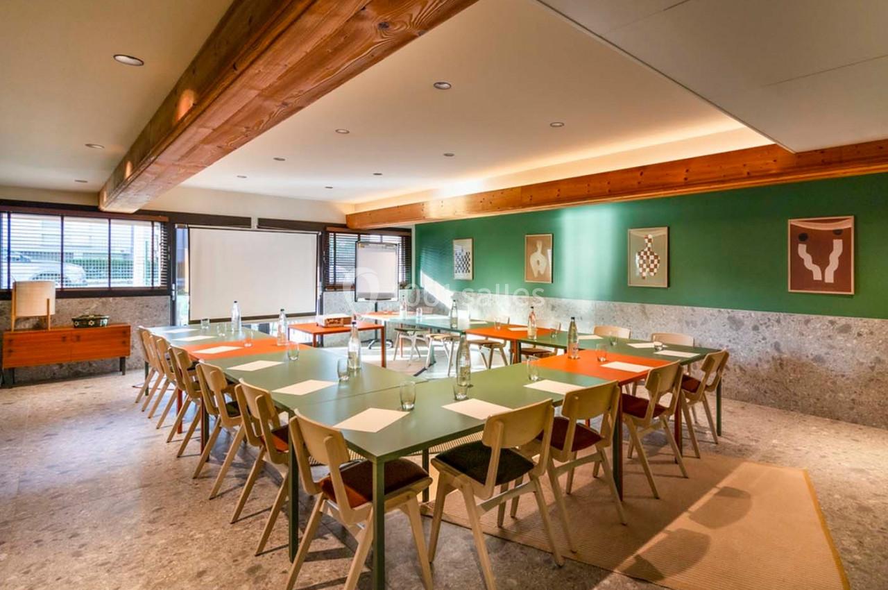 Salle de réunion lumineuse avec tables disposées en U, chaises en bois, tableau blanc et décorations murales.