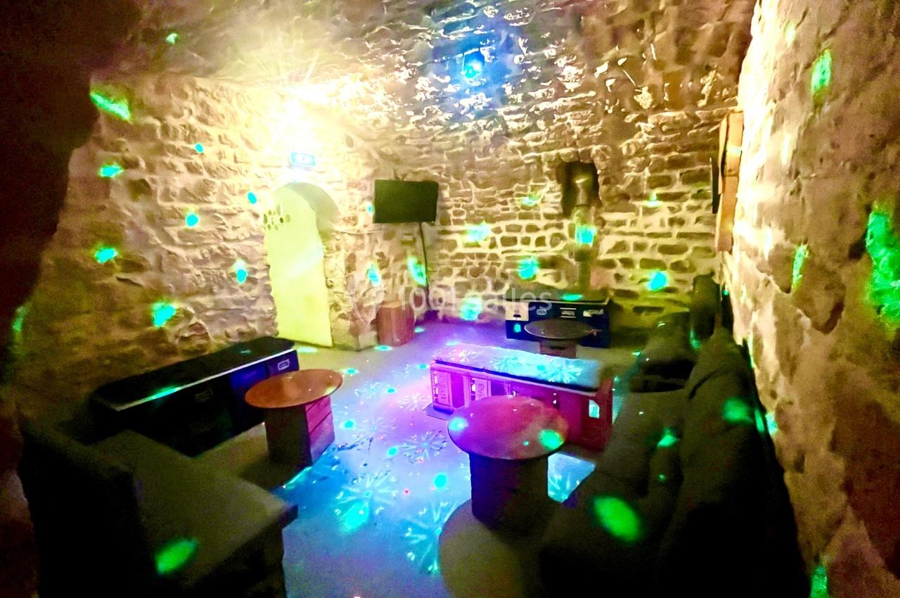 Salle voûtée en pierre éclairée par des lumières colorées, avec canapés, tables basses et écran mural.