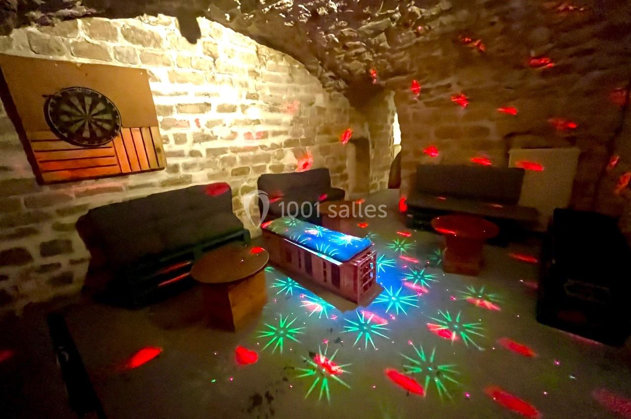 Salle voûtée en pierre avec canapés, tables basses et lumières colorées projetées au sol et sur les murs.