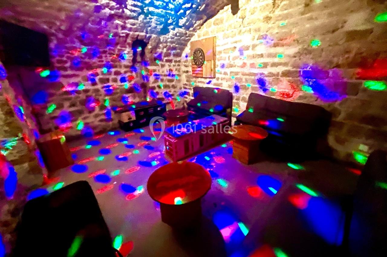 Salle voûtée en pierre éclairée par des lumières colorées, avec canapés, tables basses et cible de fléchettes au mur.