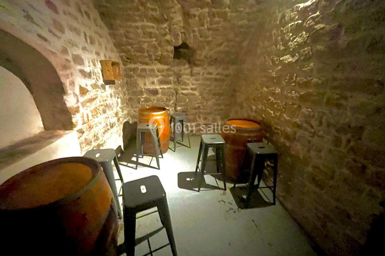 Salle voûtée en pierre avec tonneaux en bois servant de tables, entourés de tabourets noirs.