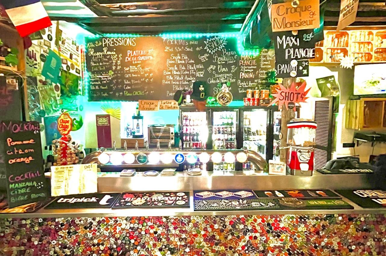 Comptoir de bar décoré de capsules de bouteilles, avec ardoises de menus, drapeaux et éclairage coloré en arrière-plan.