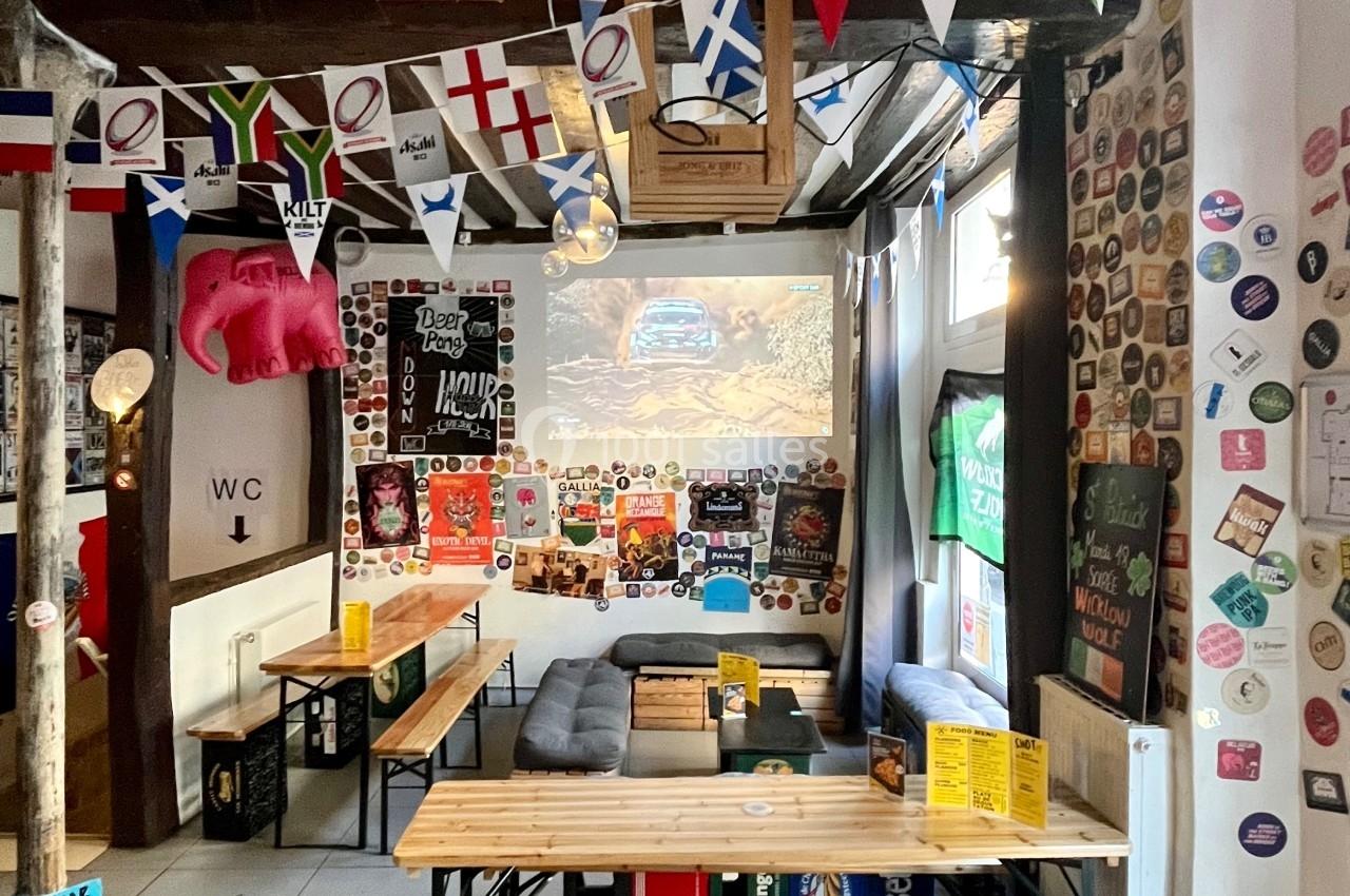 Salle conviviale décorée de fanions, affiches et objets variés, avec tables en bois et un écran projetant une image.