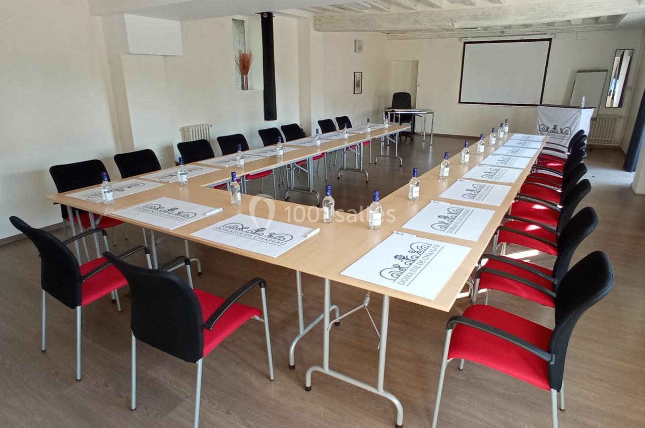 Salle de réunion lumineuse avec tables en U, chaises noires et rouges, bouteilles d'eau et écran de projection.