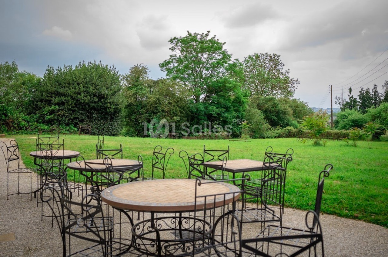 Tables et chaises en fer forgé disposées sur une terrasse donnant sur un jardin verdoyant par temps nuageux.