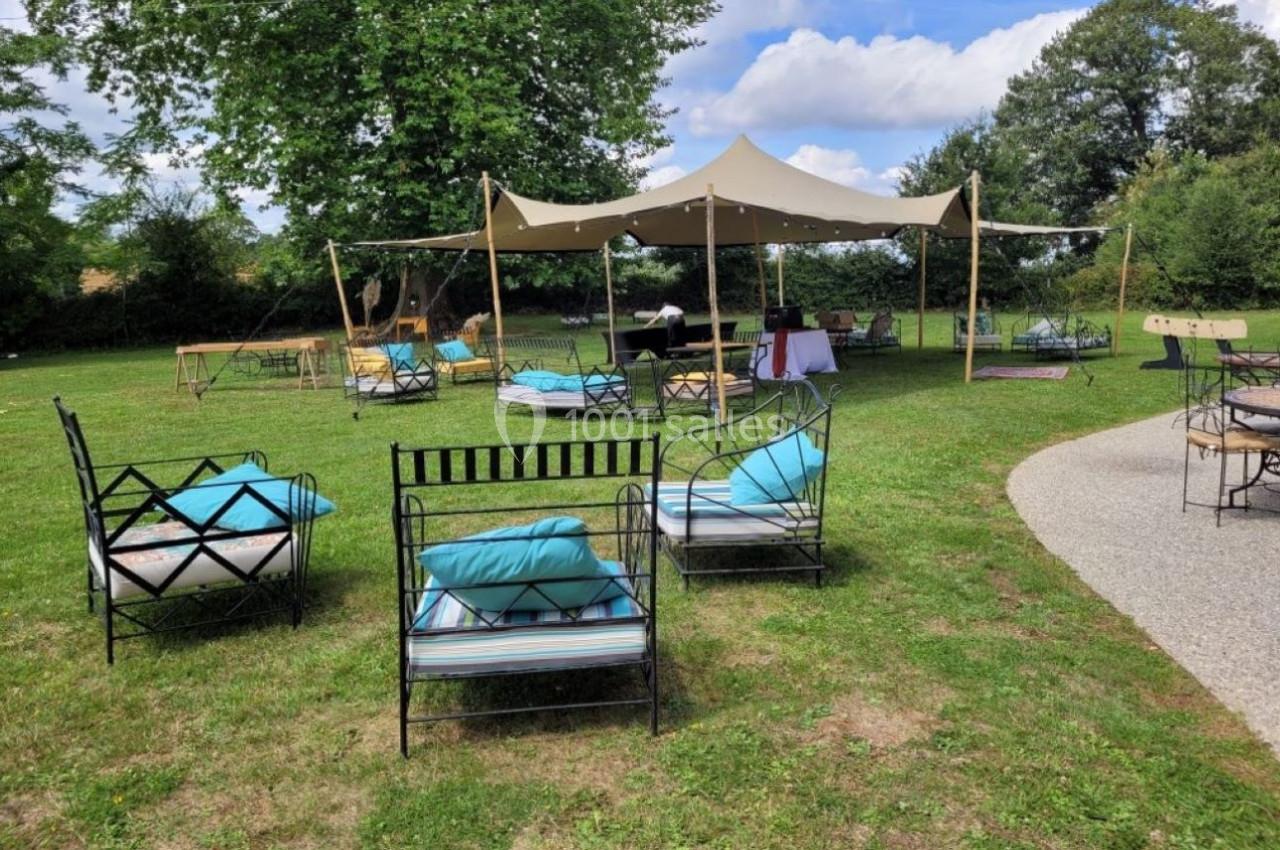 Chaises et tables disposées sur une pelouse avec un chapiteau beige en arrière-plan, entouré d'arbres.