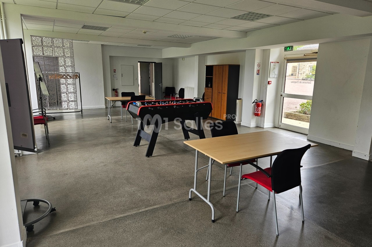 Salle lumineuse avec baby-foot, tables, chaises et sol en béton, donnant sur une sortie vitrée vers l'extérieur.