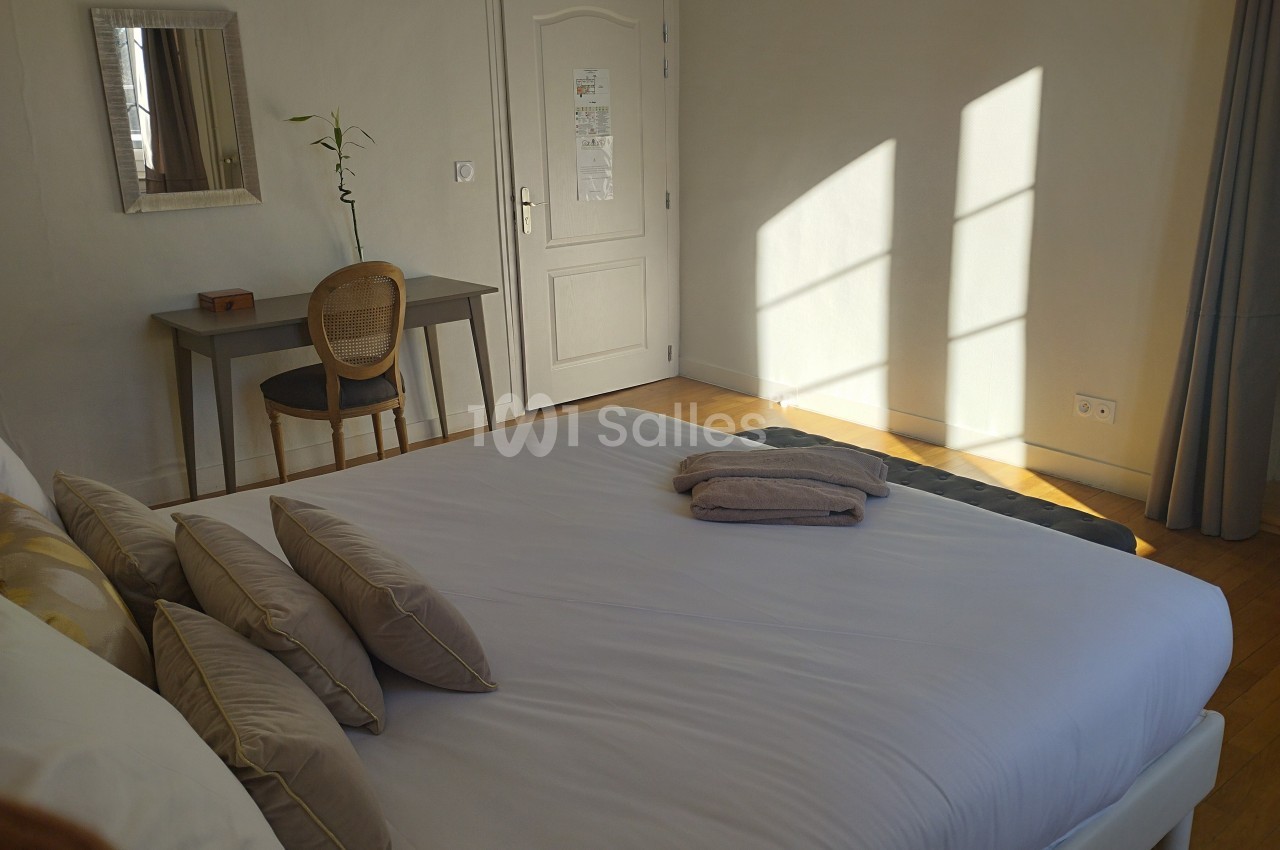 Chambre lumineuse avec un lit double, un bureau en bois clair, une chaise et une fenêtre laissant entrer la lumière du jour.