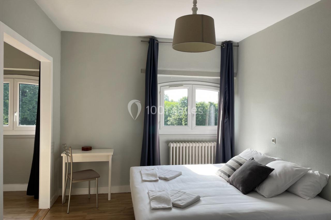 Chambre lumineuse avec lit double, bureau, fenêtre donnant sur un espace vert et rideaux sombres.