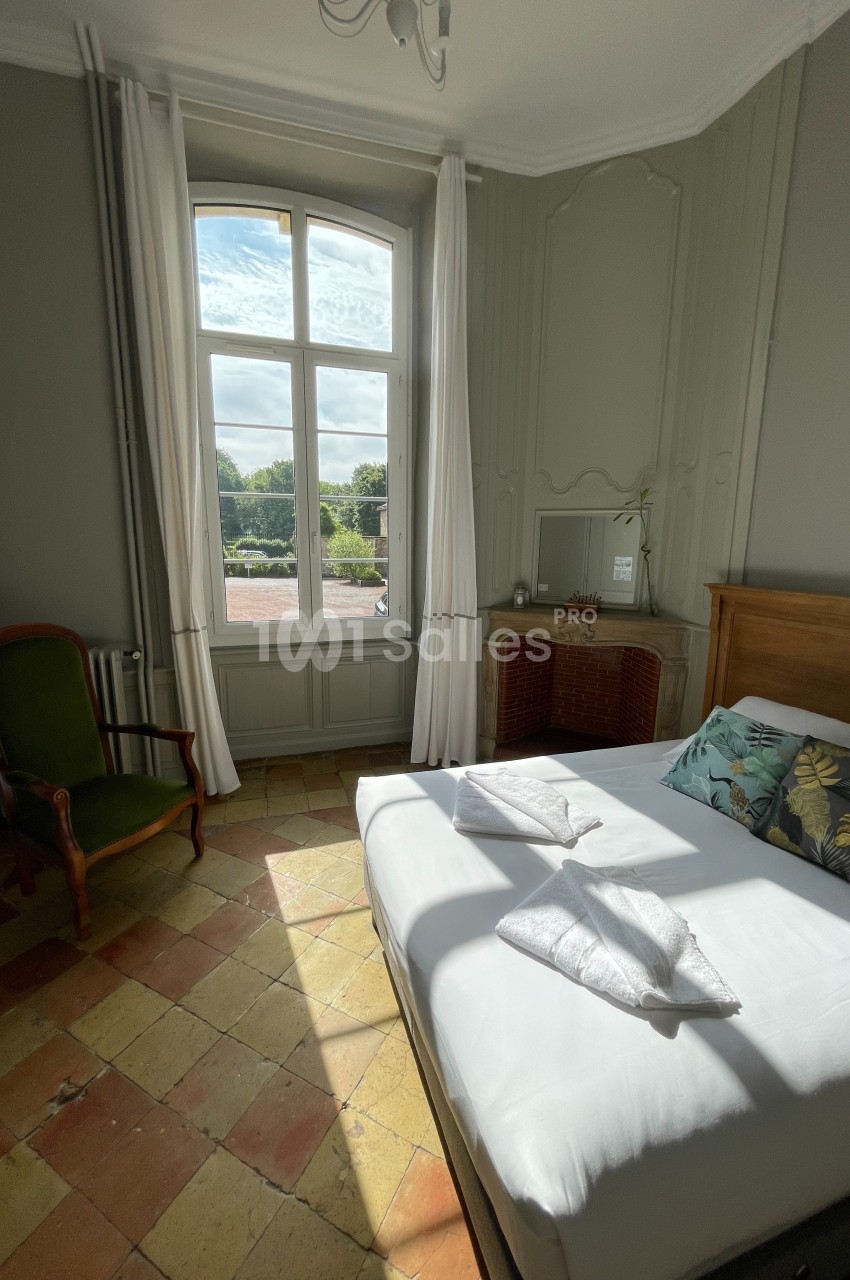 Chambre lumineuse avec un lit double, une grande fenêtre donnant sur un paysage verdoyant, et un fauteuil ancien.