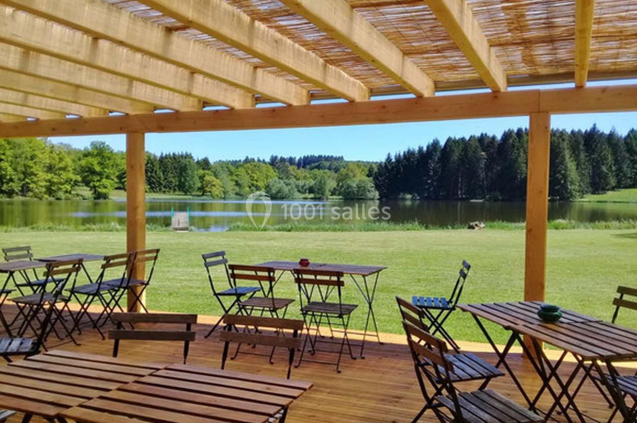 Terrasse en bois avec tables et chaises, surplombant un lac entouré de pelouse et de forêt sous un ciel dégagé.