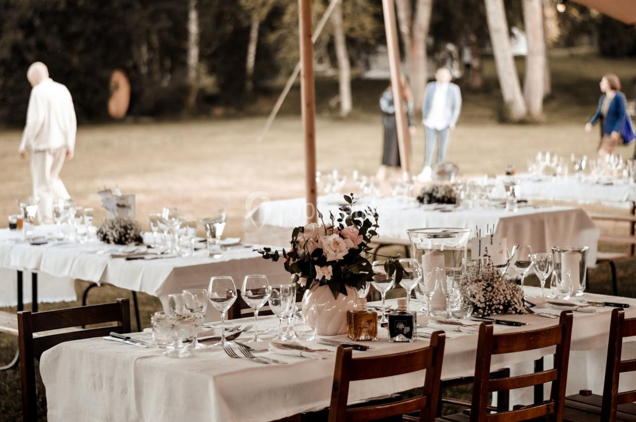 Tables dressées avec nappes blanches, vaisselle et décorations florales sous une tente en extérieur, avec des invités en…