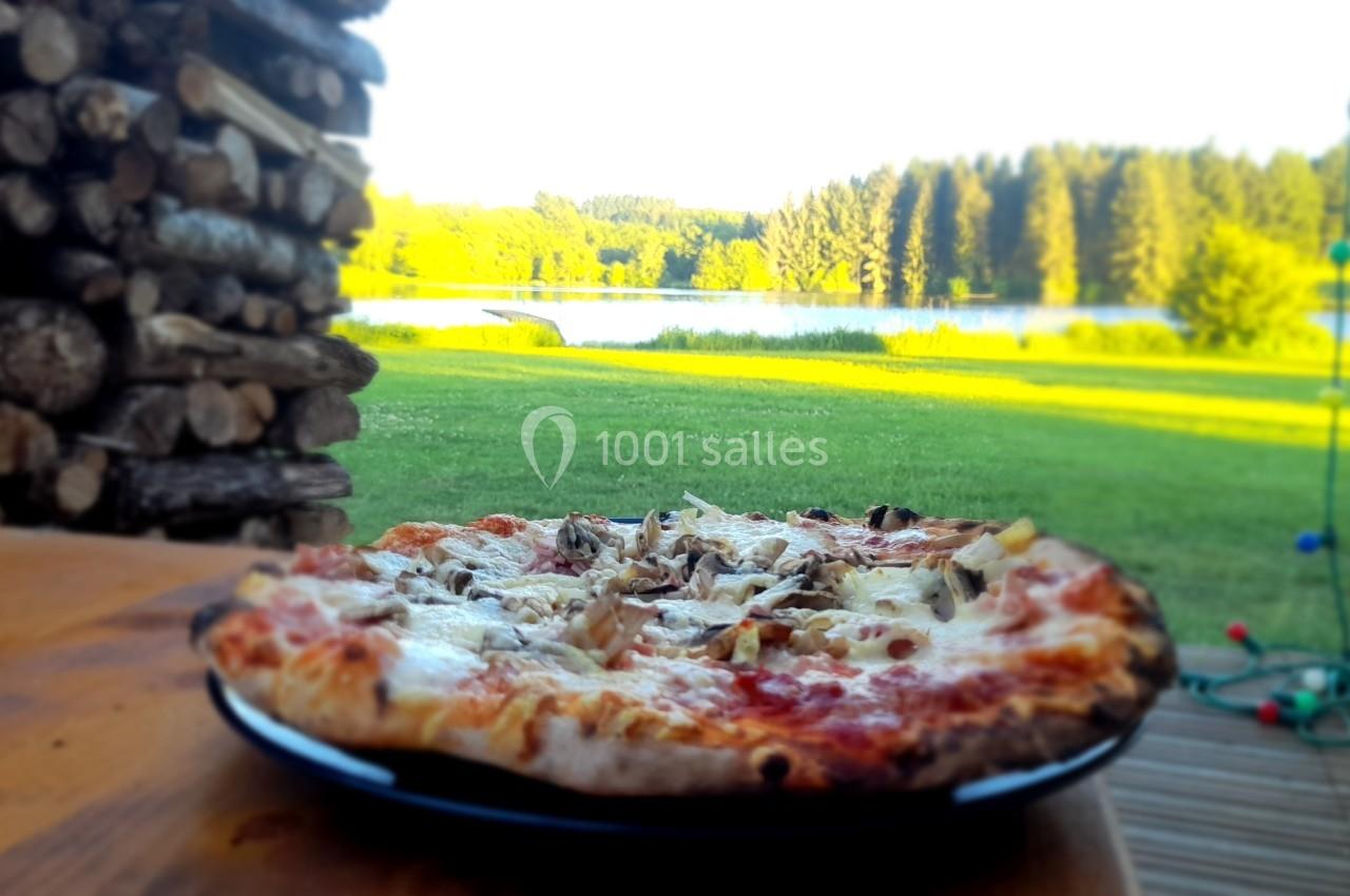 Pizza sur une assiette noire posée sur une table en bois, avec un lac et une forêt en arrière-plan.