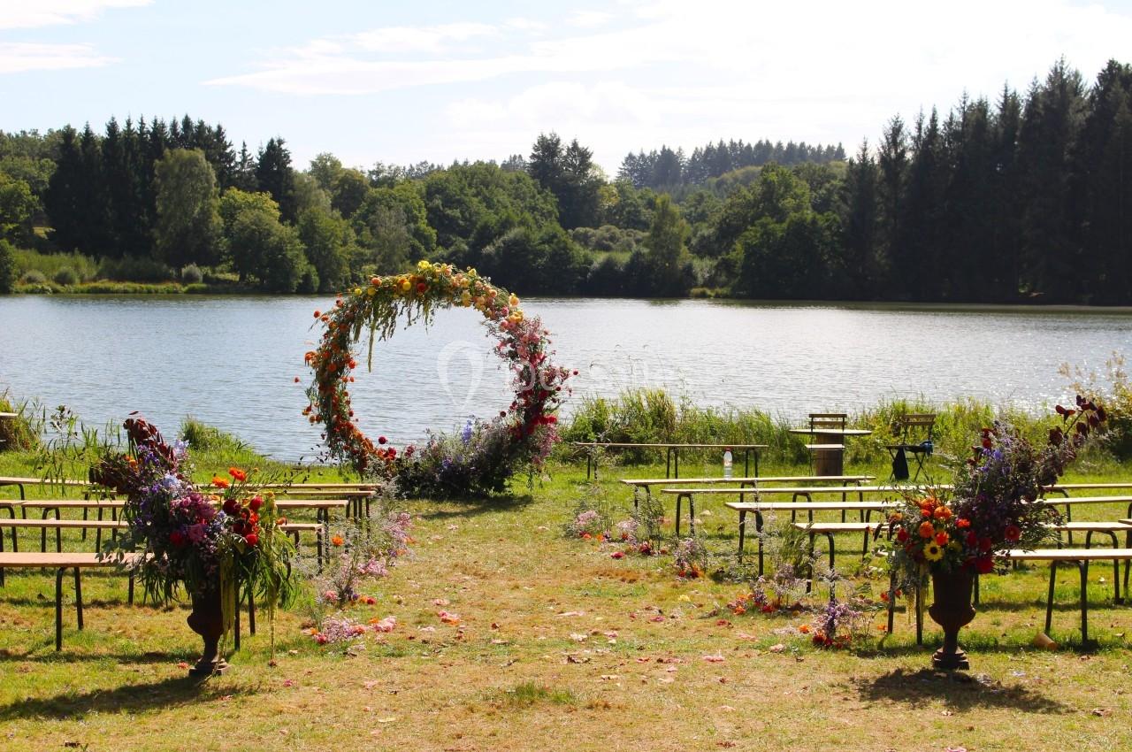 Cérémonie en plein air avec arche florale circulaire, bancs en bois et vue sur un lac entouré de forêt.