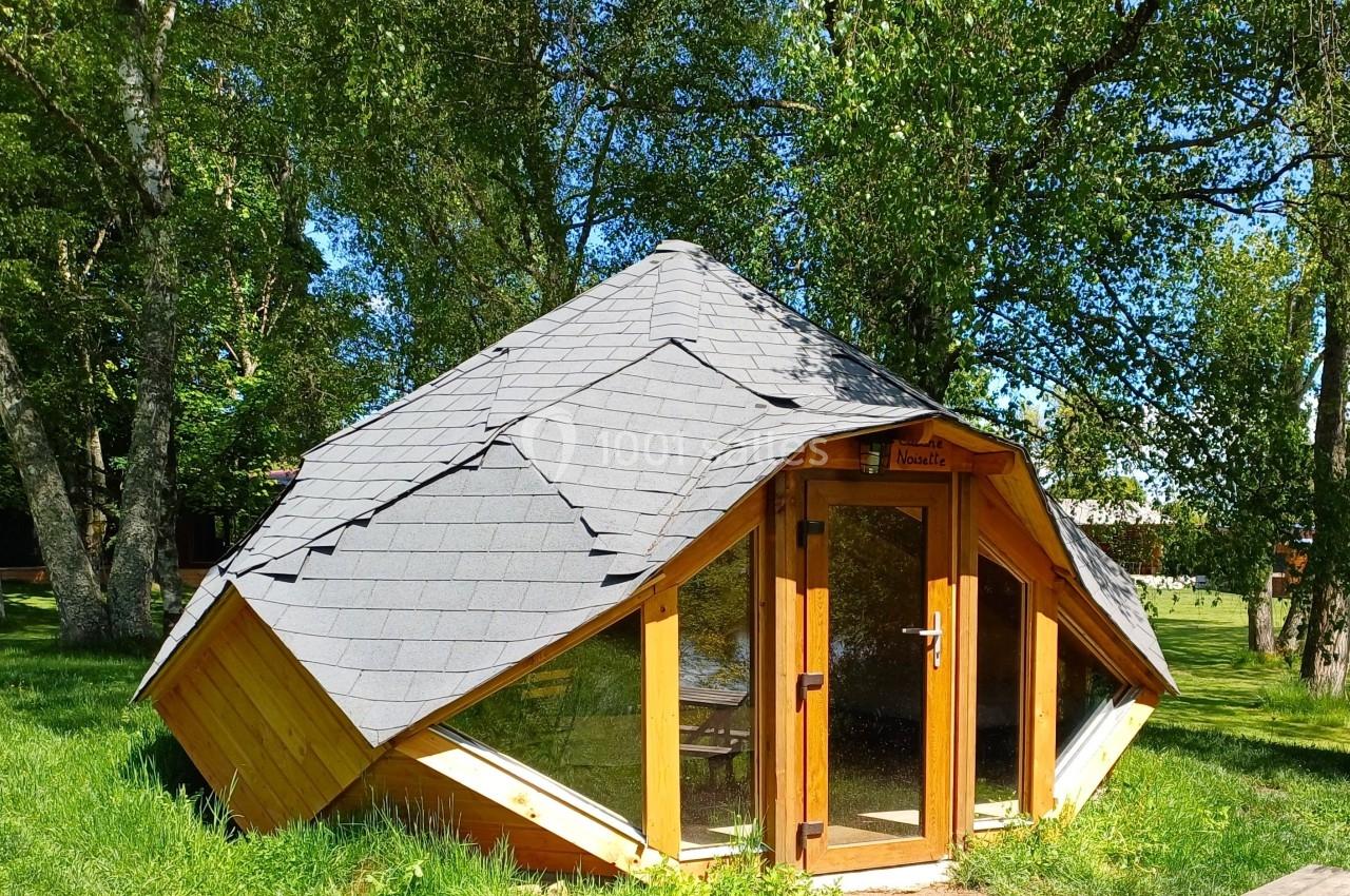 Cabane en bois au toit incliné recouvert d'ardoises, entourée d'arbres et située sur une pelouse ensoleillée.