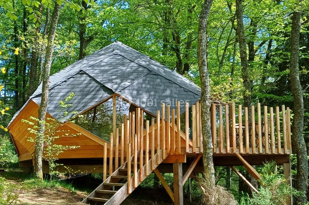 Cabane en bois sur pilotis avec terrasse, entourée d'arbres dans une forêt verdoyante.