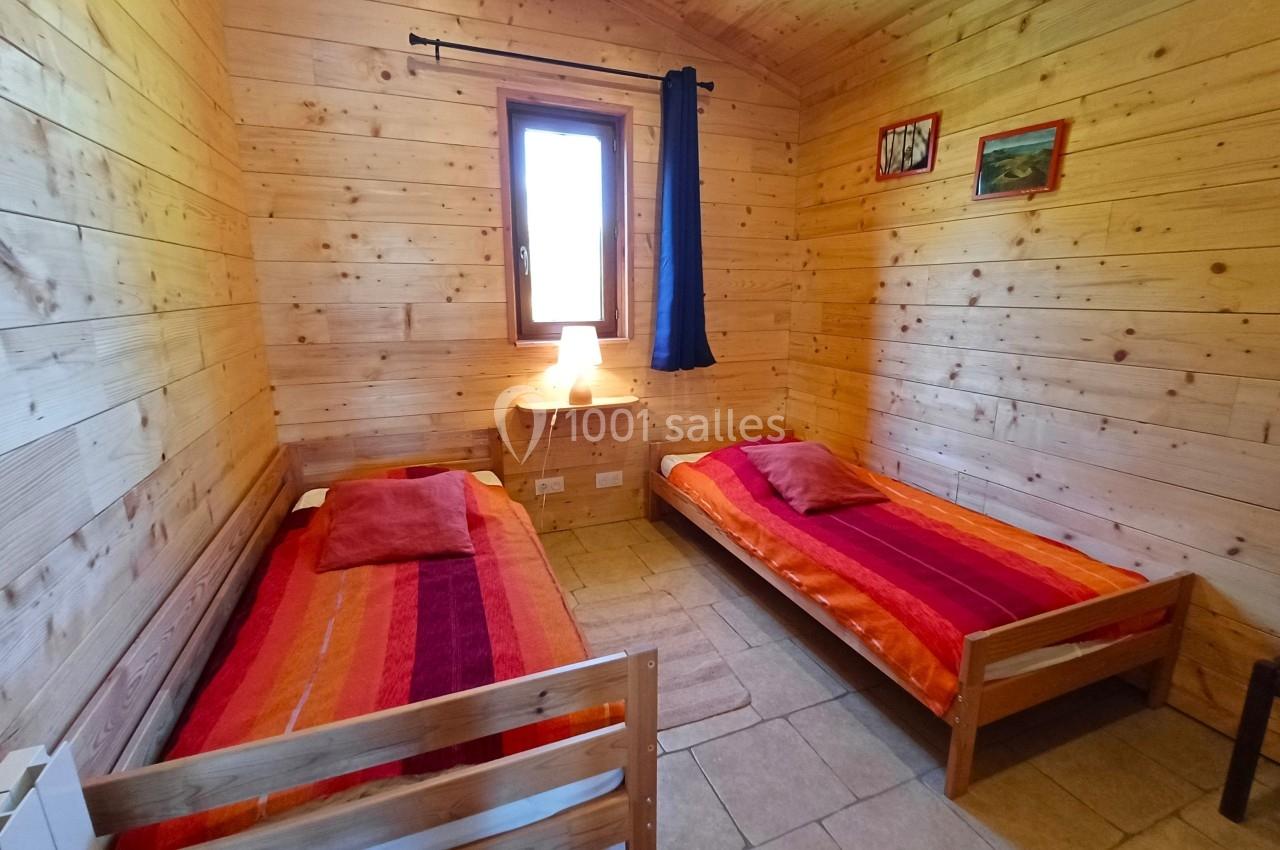 Chambre en bois avec deux lits simples, draps colorés, une fenêtre avec rideau bleu et des décorations murales.
