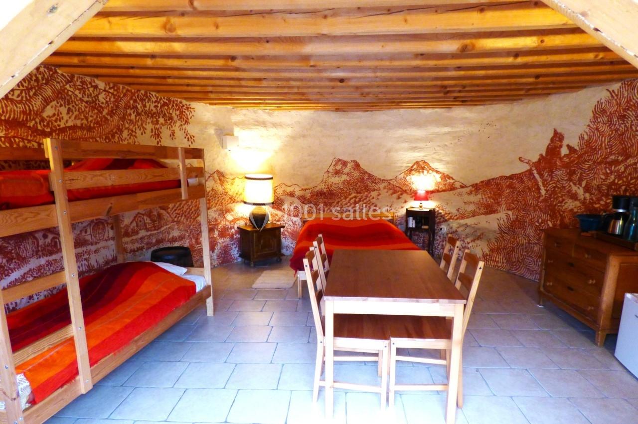 Chambre rustique avec lits superposés, lit double, table en bois et fresque murale représentant un paysage montagneux.