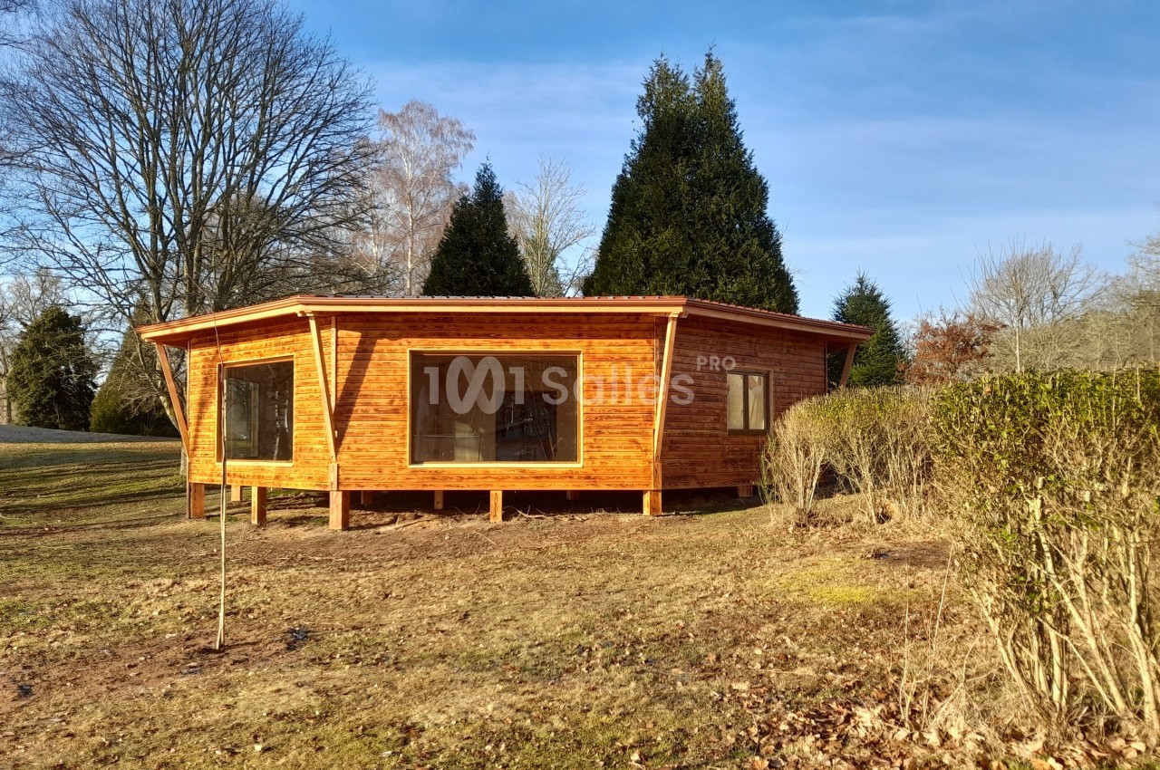 Cabane en bois avec de grandes fenêtres, située dans un espace naturel entouré d'arbres et d'arbustes.