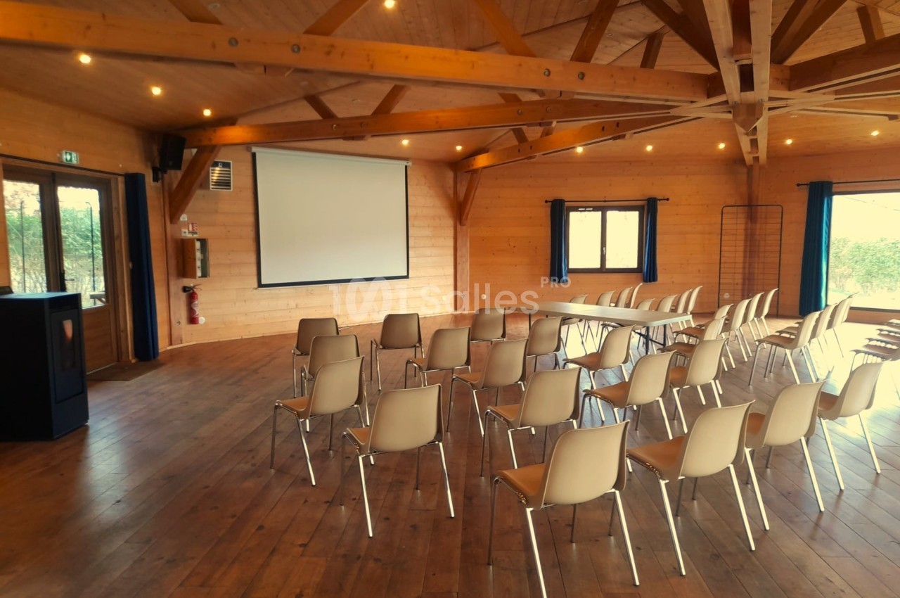 Salle en bois avec des chaises disposées face à un écran de projection, éclairée par des fenêtres et des lumières au plafond.