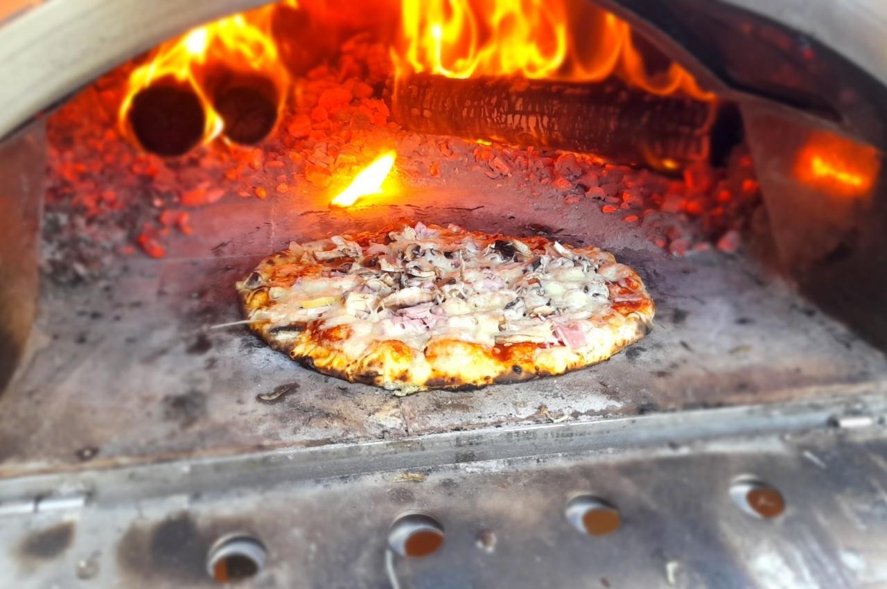 Pizza en train de cuire dans un four à bois avec des flammes et des braises visibles en arrière-plan.