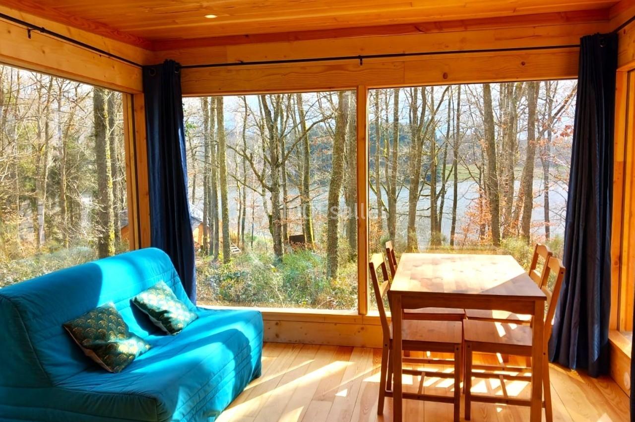 Intérieur d'une cabane en bois avec grande baie vitrée donnant sur une forêt et un lac, canapé bleu et table en bois.