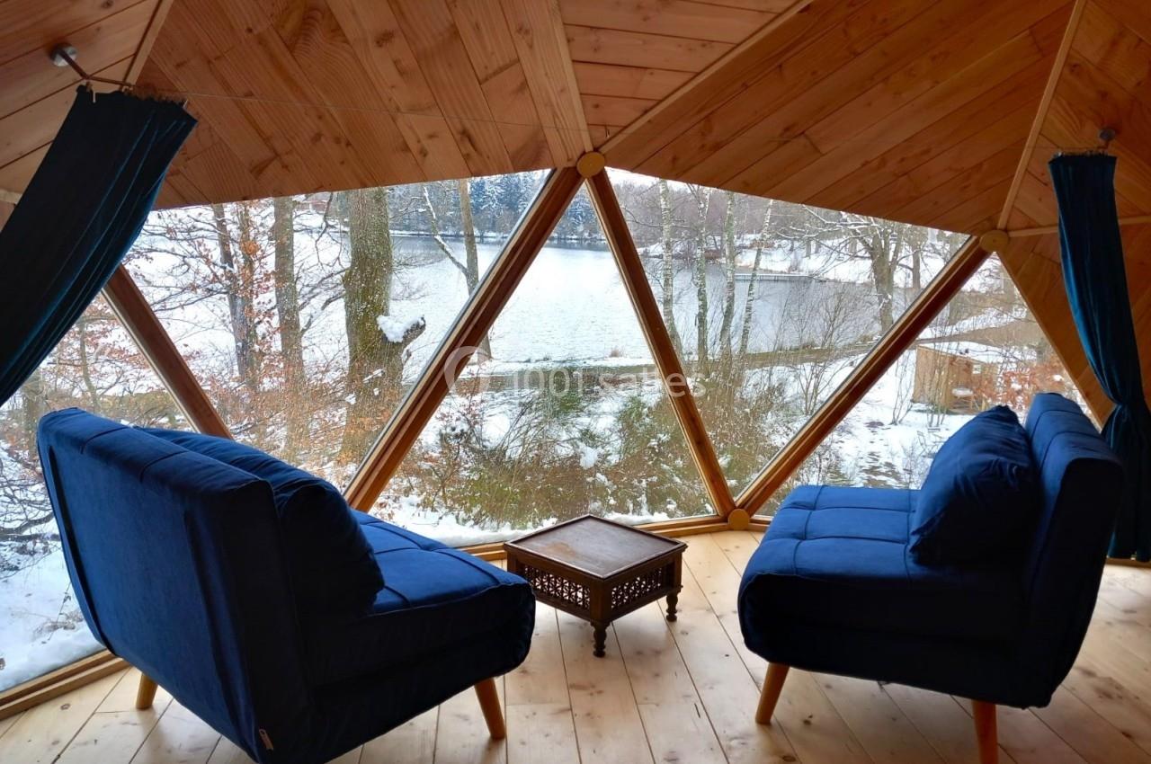 Deux fauteuils bleus et une table basse en bois dans une pièce avec vue sur un paysage enneigé à travers de grandes fenêtres.