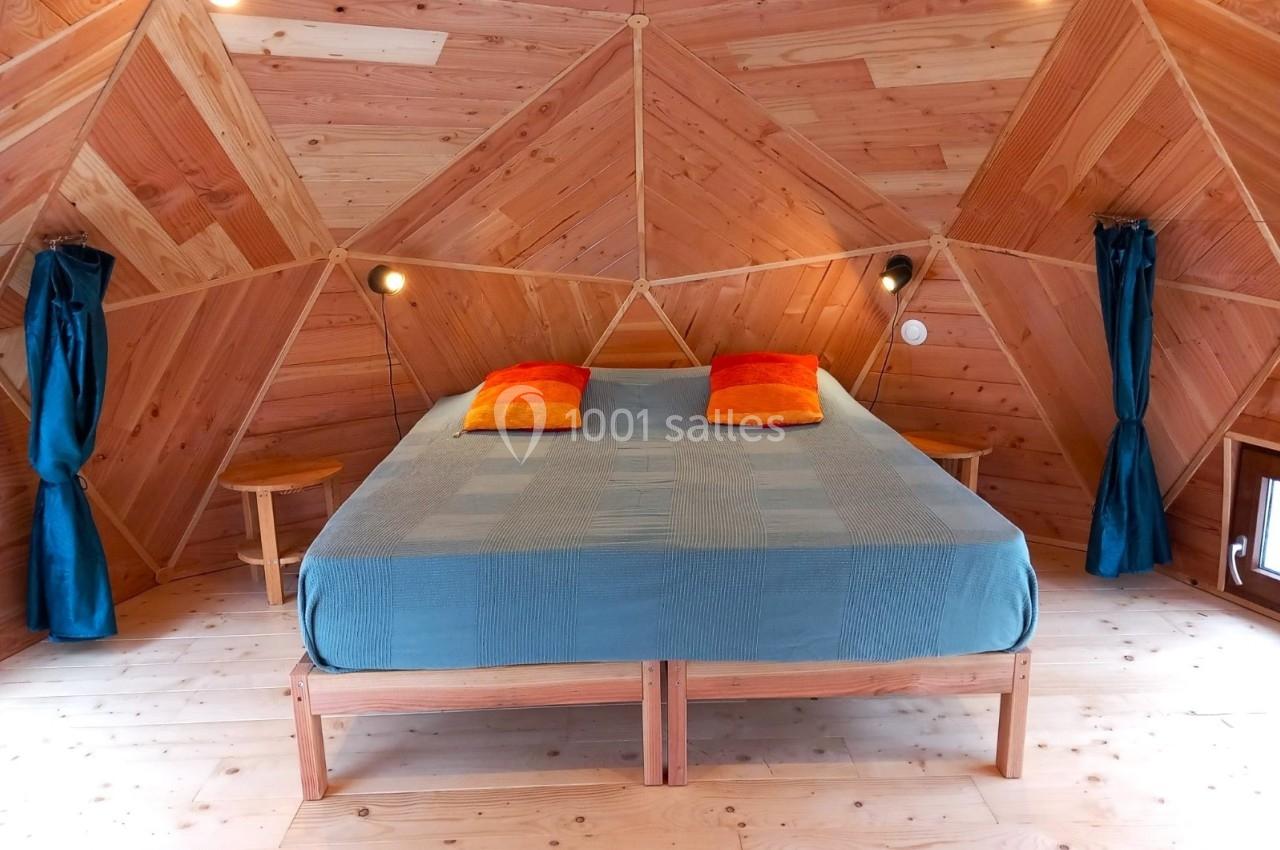 Chambre en bois avec un lit double, drap bleu, coussins orange et rideaux bleus, éclairée par des lampes murales.
