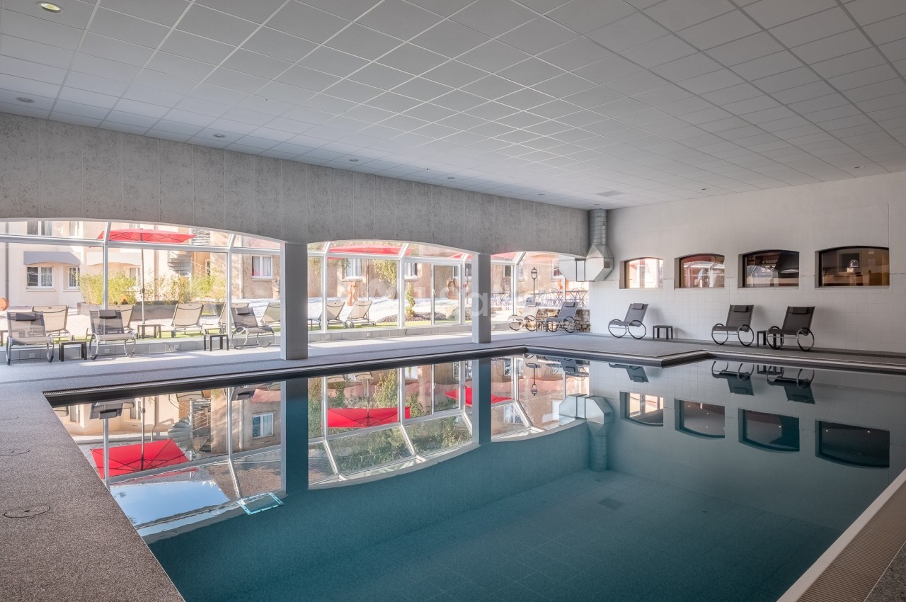 Piscine intérieure avec baies vitrées donnant sur une terrasse équipée de chaises longues et parasols rouges.