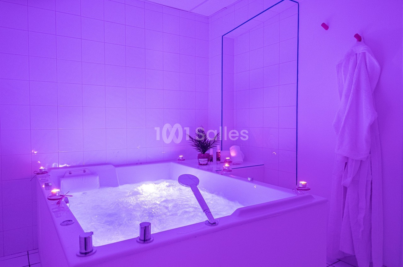 Baignoire balnéo éclairée par une lumière violette, entourée de bougies et d'un peignoir suspendu.