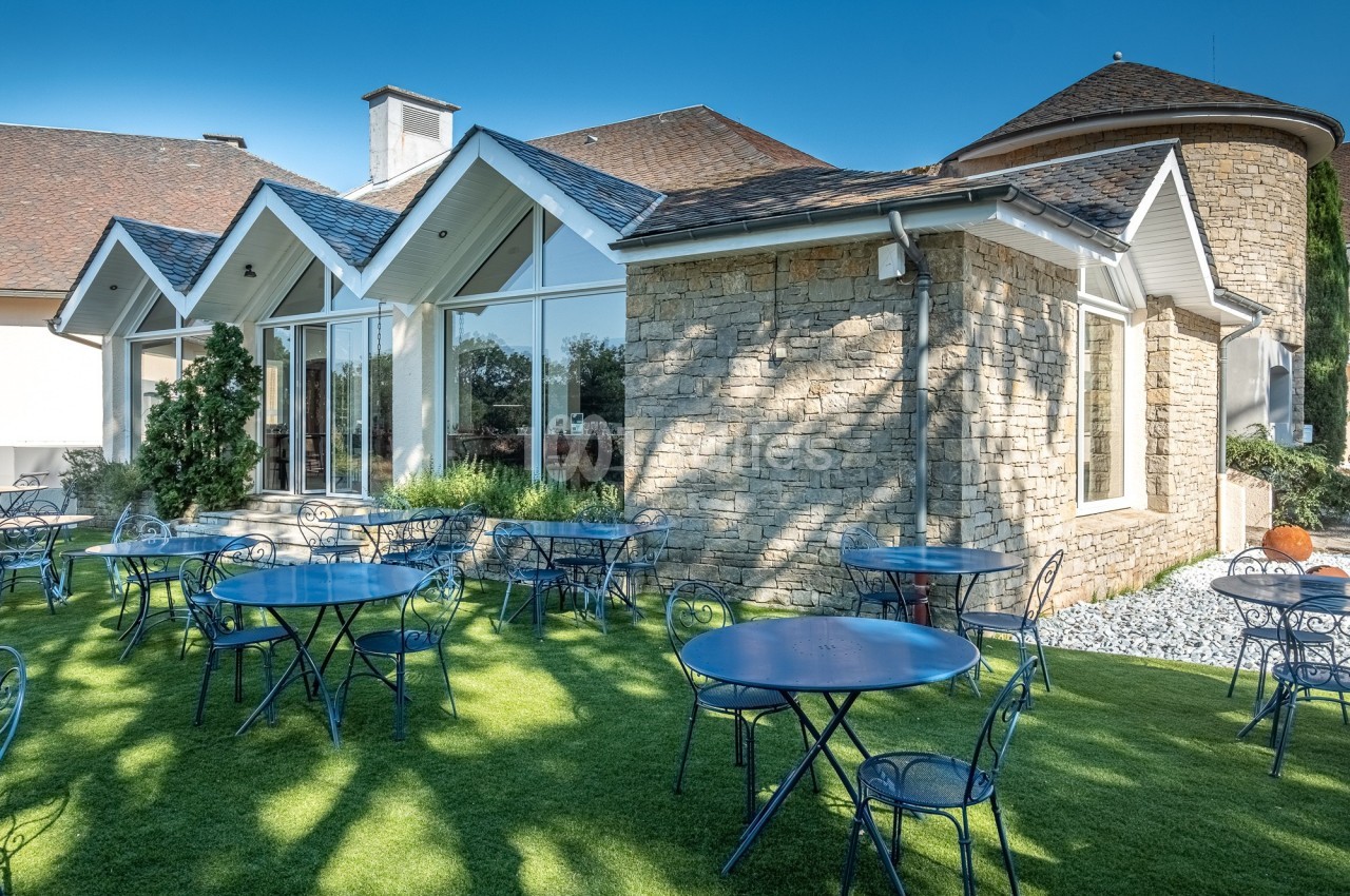 Terrasse avec tables et chaises en métal bleu sur une pelouse, devant un bâtiment en pierre avec de grandes fenêtres.
